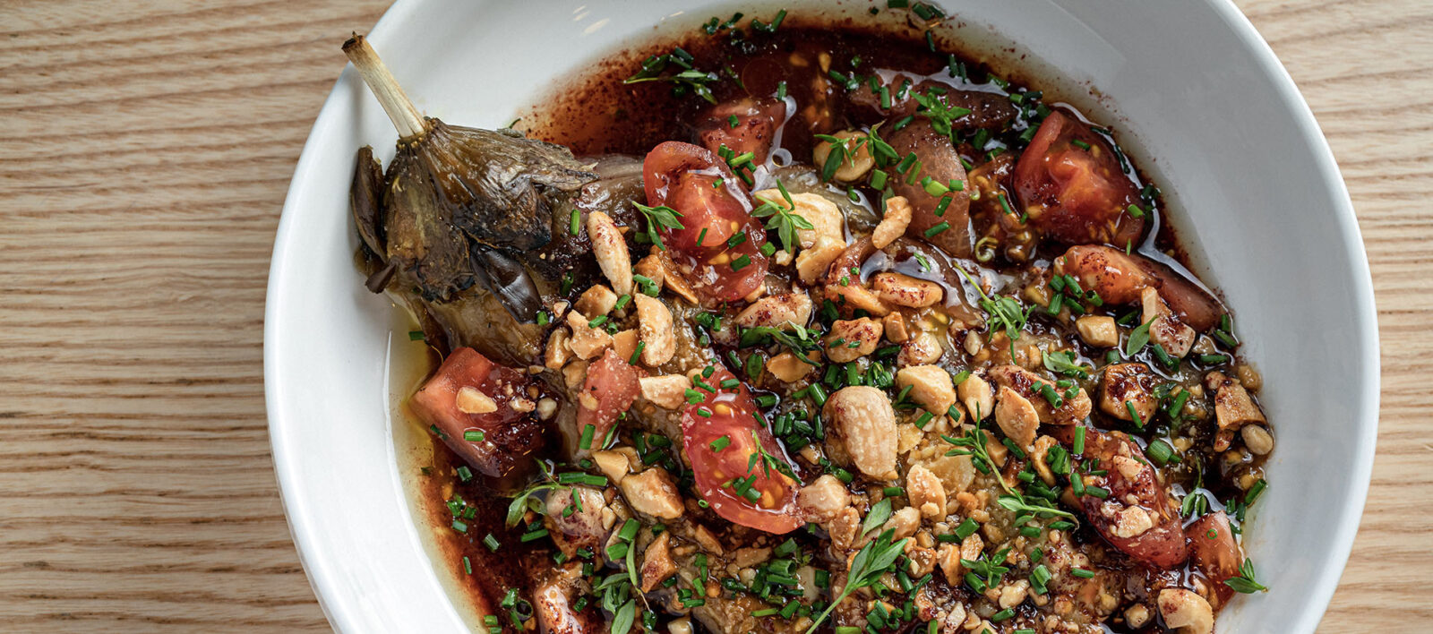 Whole fish head in a spicy tomato‑peanut broth, garnished with chopped herbs and green onions in a white bowl.